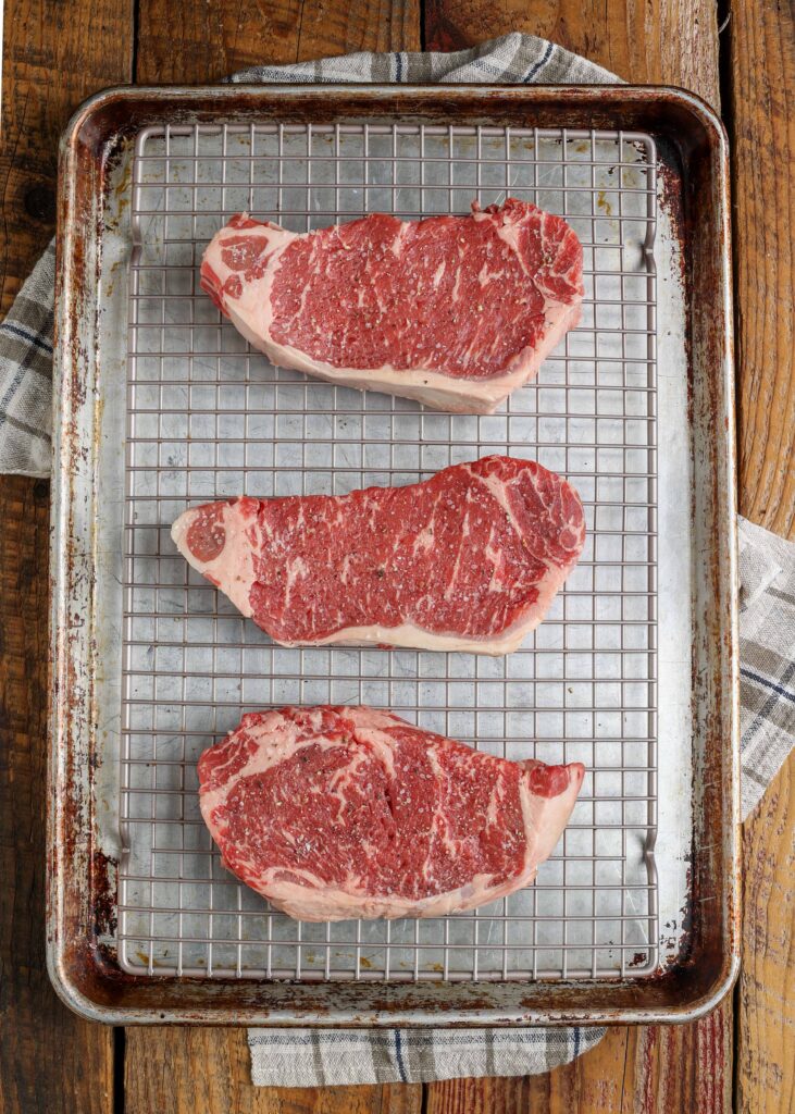 raw marbled New York strip steaks on rack over sheet pan