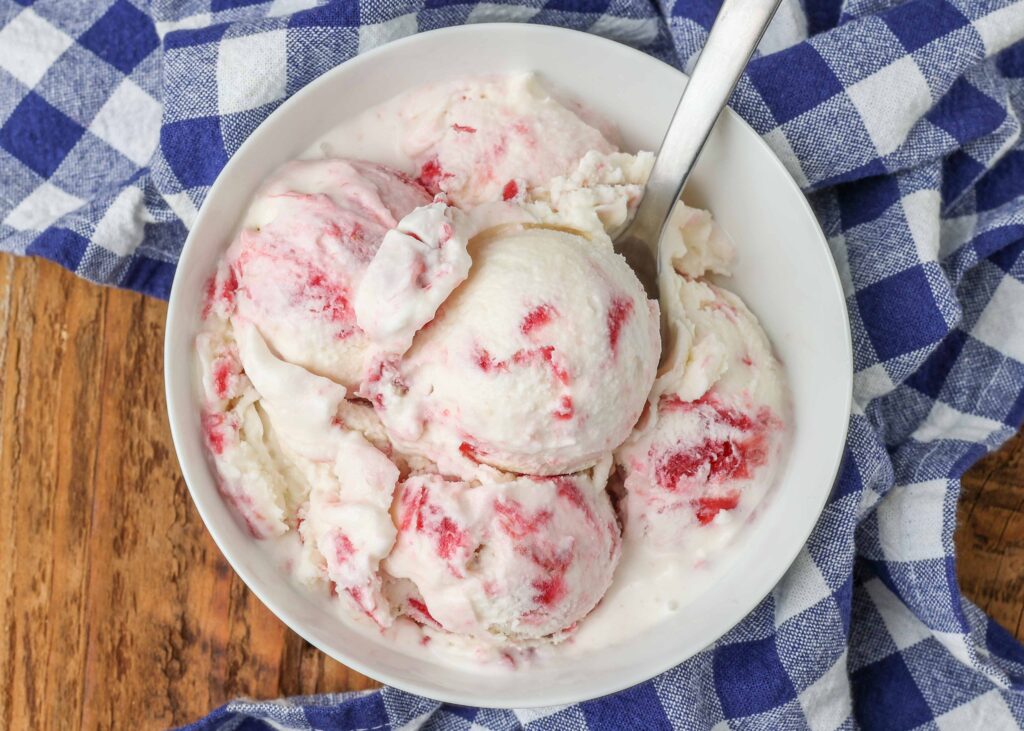 Overhead horizontal shot of white and red rhubarb swirl ice cream, served in a white bowl with a silver spoon and a checkered blue and white towel